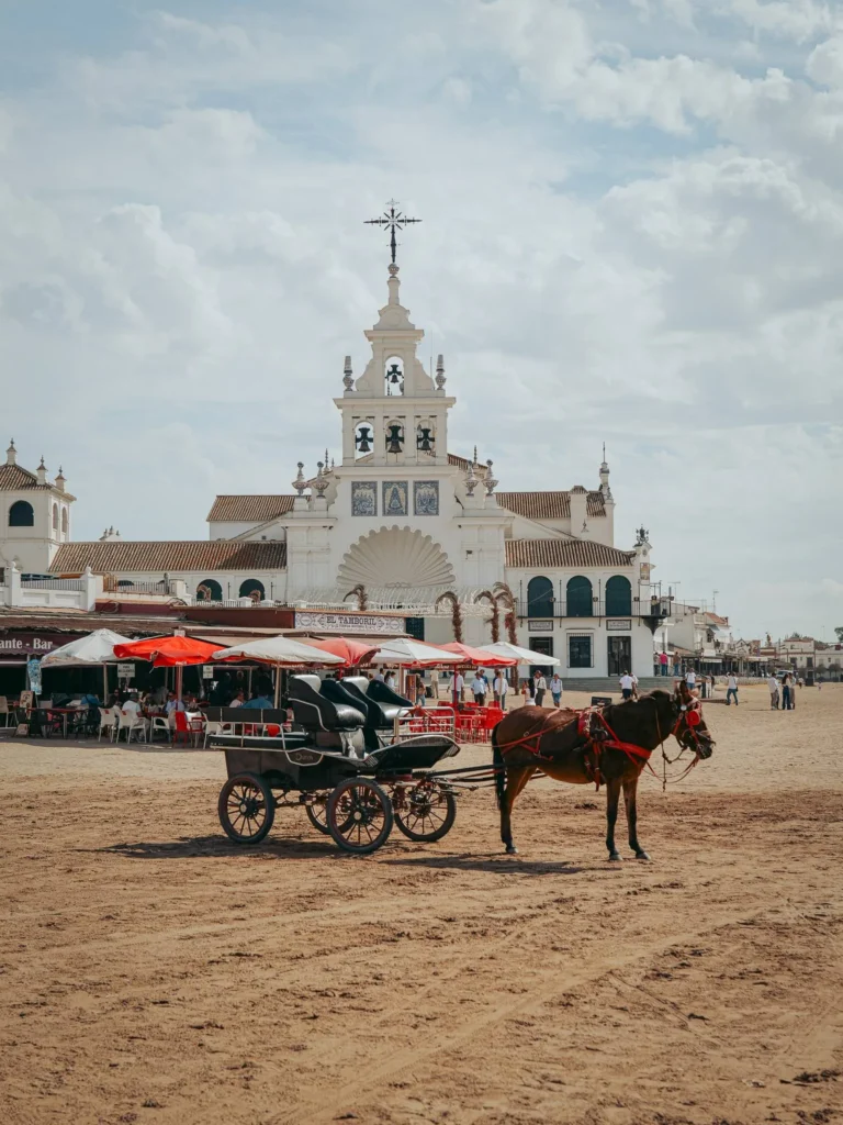 Coche de caballos sobre la arena con el santuario del Rocío y terrazas al fondo