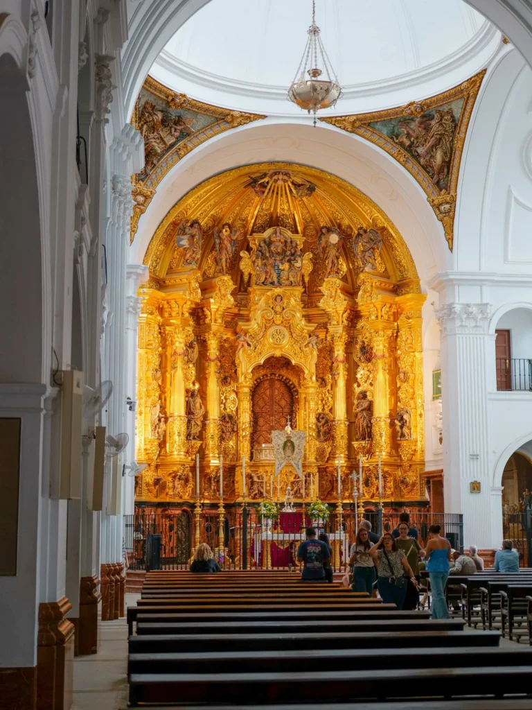 Retablo dorado e interior blanco del santuario del Rocío con visitantes junto a los bancos